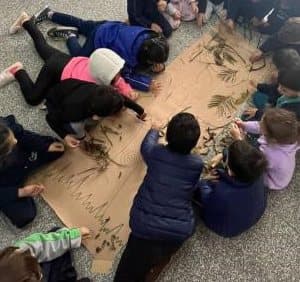 Crianças da Educação Infantil montando, no chão, um cartaz coletivo com as partes de uma árvore, usando folhas secas e gravetos.