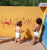 Duas crianças pequenas, de fralda, pintam com tinta vermelha um muro amarelo do solário.