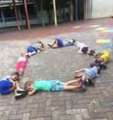 Crianças deitadas de bruços no chão de lajotas do pátio, formando uma roda.