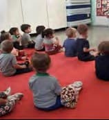 Crianças sentadas sobre um tapete vermelho na sala de aula, vistas de costas.