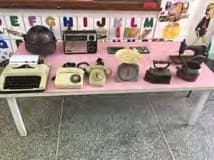 Objetos antigos expostos sobre uma mesa rosa: máquina de escrever, telefones, rádio, ferros de passar e panelas.