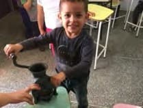 Menino sorridente gira a manivela de um moedor antigo apoiado numa mesa da sala.
