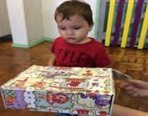 Bebê de camiseta vermelha olha para uma caixa estampada e fechada à sua frente.