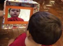 Uma criança, de costas, abre a caixa, e o rosto de um bebê aparece no espelho de moldura laranja dentro da tampa.