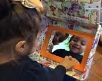 Duas crianças olham juntas o espelho dentro da caixa aberta e sorriem para o próprio reflexo.