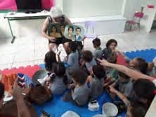 Crianças sentadas no tatame olham para a professora, que mostra uma foto antiga de família.