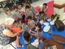 Roda de crianças no tatame com a professora, em volta de pratos, louças, cestos e outros objetos.