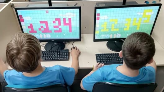 Dois alunos em computadores construindo imagens pixeladas coloridas na tela.