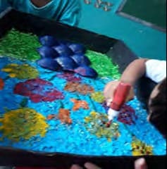 Mão de criança, com um bastão de cola colorida, sobre um painel coberto de sementes, papéis picados e formas em azul, verde, vermelho e amarelo.