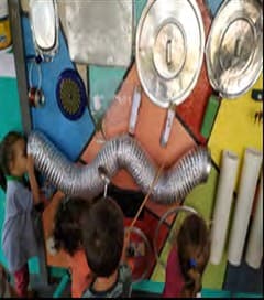 Crianças de costas diante de uma parede sonora colorida, com tampas de panela, mangueira sanfonada, canos e outros objetos presos nela.
