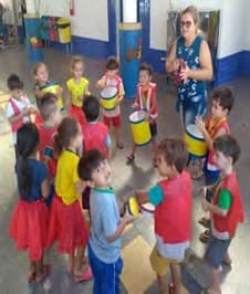Crianças em roda no pátio tocam tambores feitos de latas, com baquetas, junto da professora.