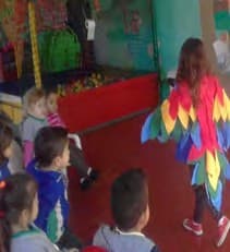 Uma criança fantasiada de arara, com asas de penas coloridas, diante de colegas sentados que assistem à cena.