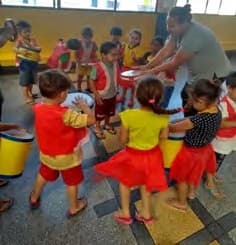 Crianças caracterizadas, com saias de babados vermelhas e amarelas, brincam em roda com a professora.