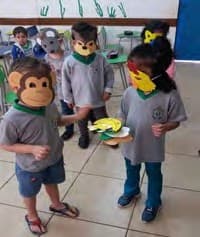 Crianças com máscaras de macaco e de outros bichos feitas de papel representam uma cena na sala.
