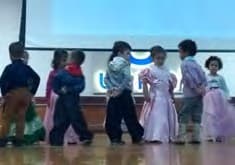 Crianças com trajes de época, vestidos longos e casacos, dançam em pares num palco.