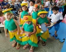 Crianças fantasiadas com saias de franjas verdes e amarelas e chapéus de frutas dançam no pátio.
