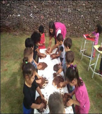 No gramado, crianças em volta de uma mesa coberta de papel branco modelam montes de terra com as mãos, com a professora ao fundo.