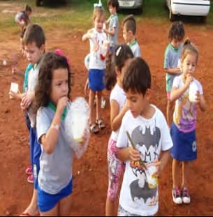 Crianças de uniforme, num terreno de terra vermelha, sopram bolhas de sabão com canudos e copinhos.