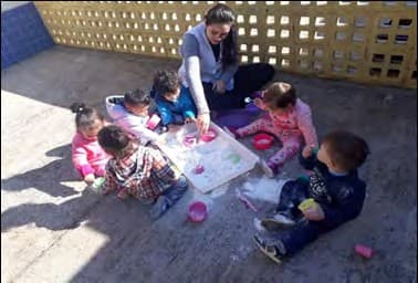 Bebês sentados no chão do pátio, ao sol, com a professora, mexem com as mãos numa massa branca espalhada sobre papel e em potinhos coloridos.