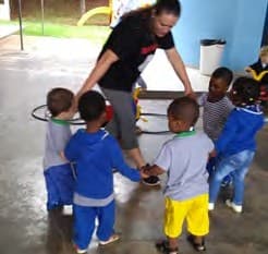 Professora curvada, de mãos dadas com crianças pequenas numa roda, numa área coberta.