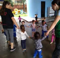 Duas professoras e um grupo de crianças pequenas brincam de roda num pátio coberto, com brinquedos ao fundo.