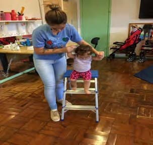 Professora segura as mãos de um bebê que se equilibra de pé sobre um banquinho.