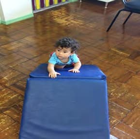 Bebê engatinha sobre um colchonete azul dobrado em rampa, no chão de tacos.