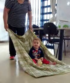 Professora puxa uma coberta xadrez com um bebê sentado sobre ela.