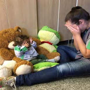 Professora cobre o rosto com as mãos diante de um bebê sentado entre bichos de pelúcia, que também cobre os olhos.