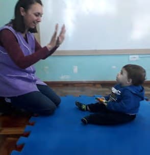 Professora sentada no chão bate palmas e canta para um bebê sentado à sua frente, num tatame azul.
