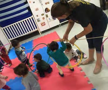 Professora segura bambolês de pé enquanto crianças passam por dentro deles, sobre um tatame colorido.