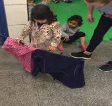 Menina estende um tecido num varal, ao lado de outras crianças, diante de uma parede verde.