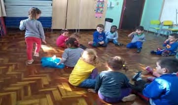 Crianças sentadas em roda no chão da sala; uma menina caminha por fora da roda.
