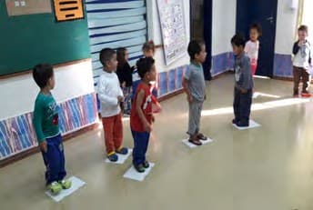 Crianças em pé, cada uma sobre uma folha marcada no chão da sala.
