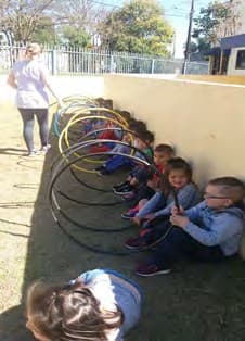 Crianças sentadas junto a um muro seguram bambolês formando um túnel, com uma professora ao fundo.