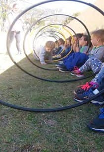 Túnel de bambolês segurados por crianças sentadas na grama, visto de uma das pontas.