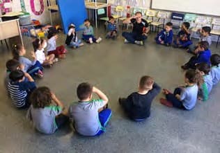 Crianças sentadas em roda no chão da sala, com a professora, cantando e fazendo gestos.