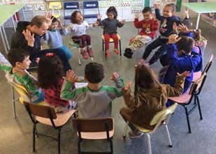 Crianças sentadas em cadeiras, em roda, erguem os braços acompanhando a história contada pelo professor.