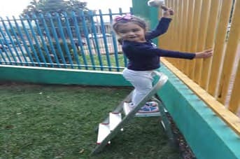 Menina sorridente sobe uma escada de metal encostada num muro, no pátio gramado.