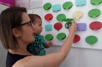 Professora com um bebê no colo aponta, num painel branco, balões de fala coloridos com fotos das crianças.