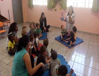 Professora de pé conta uma história com fantoches para crianças sentadas no chão da sala.