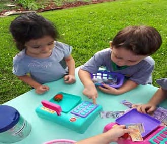 Duas crianças brincam de mercado numa mesa ao ar livre, com caixa registradora, cestinha e dinheiro de brinquedo.