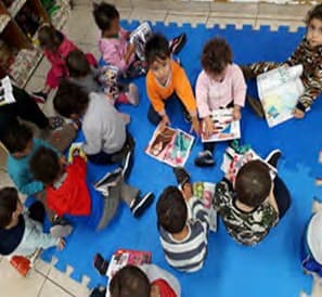 Crianças pequenas sentadas no tatame azul folheiam livros e revistas.