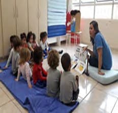 Crianças sentadas no colchonete ouvem a professora, que mostra um livro aberto.