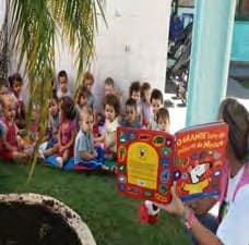 No pátio, crianças sentadas na grama olham a professora, que lê para elas um livro grande e colorido.