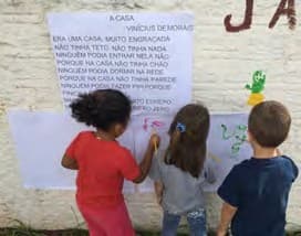 Três crianças de costas desenham num papel colado no muro, abaixo do cartaz com a letra da música «A casa», de Vinicius de Moraes.