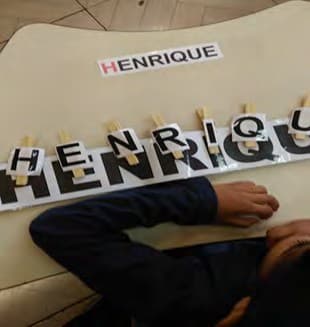 Sobre a mesa, o crachá com o nome Henrique e as letras do nome presas com prendedores em cima de uma tira com o nome escrito.