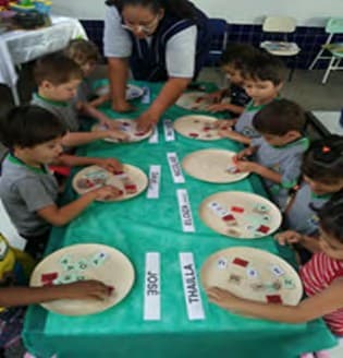 Crianças em volta de uma mesa, cada uma diante de um prato com letras e de uma tira com o próprio nome, com a professora ao fundo.