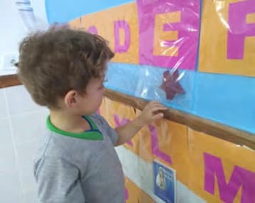 Menino mexe num saquinho com objetos preso abaixo das letras do alfabeto concreto na parede.