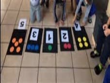 No chão da sala, tapetes pretos com os números 5, 3, 1, 2 e 4, cada um com a mesma quantidade de tampinhas coloridas; em volta, os pés das crianças.